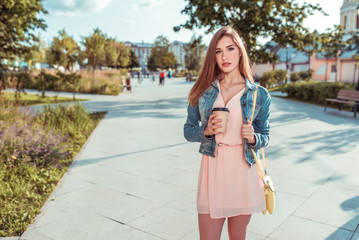 beautiful girl student standing city summer, holding cup coffee tea, yellow handbag, waiting for friends girlfriends, long hair, tanned slim figure. Pink dress casual makeup. Free space for text.