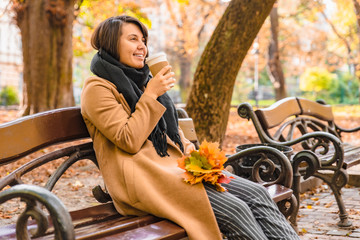 woman sitting on the bench at autumn city park drinking coffee