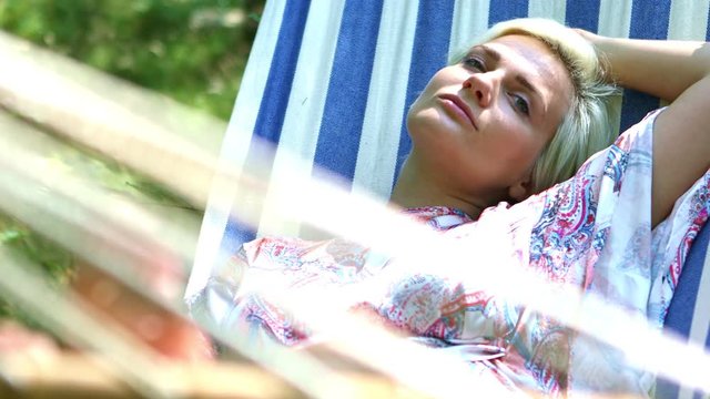 Blonde woman resting on a blue and white hammock during daytime wearing summer clothes with trees and grass as background