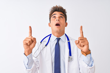 Young handsome doctor man wearing stethoscope over isolated white background amazed and surprised looking up and pointing with fingers and raised arms.