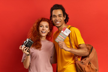 Happy triumphing diverse couple rejoices having trip soon, holding passport and flying tickets, holds the old film camera and orange backpack, poses against red wall. Vacation concept