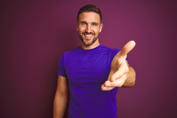 Fototapeta premium Young man wearing casual purple t-shirt over lilac isolated background smiling friendly offering handshake as greeting and welcoming. Successful business.