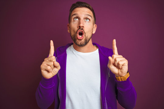 Young fitness man wearing casual sports sweatshirt over purple isolated background amazed and surprised looking up and pointing with fingers and raised arms.
