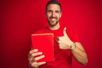 Young man using digital touchpad tablet over red isolated background happy with big smile doing ok sign, thumb up with fingers, excellent sign