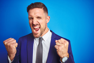 Close up picture of young handsome business man over blue isolated background very happy and excited doing winner gesture with arms raised, smiling and screaming for success. Celebration concept.