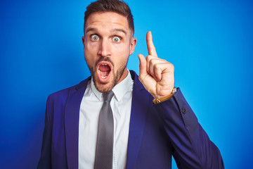 Close up picture of young handsome business man over blue isolated background pointing finger up with successful idea. Exited and happy. Number one.