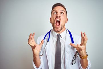 Young handsome doctor man wearing white professional coat over isolated background crazy and mad...