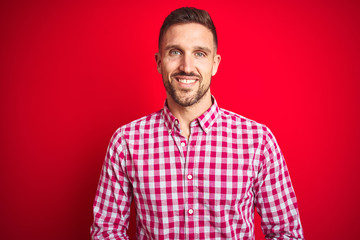 Fototapeta premium Young handsome man over red isolated background with hands together and crossed fingers smiling relaxed and cheerful. Success and optimistic