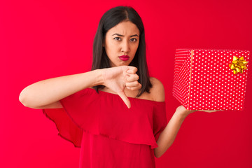 Young beautiful chinese woman holding birthday gift standing over isolated red background with...