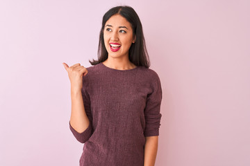 Young chinese woman wearing purple sweater standing over isolated pink background smiling with happy face looking and pointing to the side with thumb up.