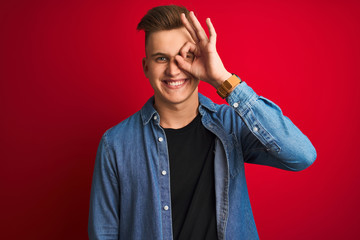 Obraz premium Young handsome man wearing denim shirt standing over isolated red background doing ok gesture with hand smiling, eye looking through fingers with happy face.