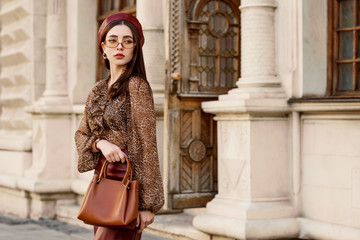 Outdoor fashion portrait of young elegant, luxury lady wearing faux leather beret, skirt, leopard print blouse, beige glasses, holding brown bag, posing in street of European city. Copy, empty space