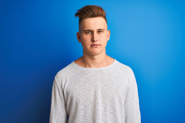 Young handsome man wearing white casual t-shirt standing over isolated blue background with a confident expression on smart face thinking serious