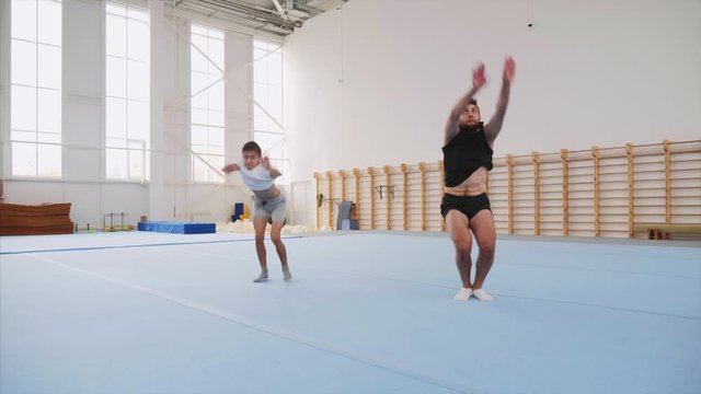 A cartwheel and a tripple back flip are made by a male sportman and a teen boy synchronously on the blue mats at sport centre, steadicam shot, back view, in slow motion.