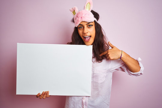 Young woman wearing pajama and sleep mask holding banner over isolated pink background with surprise face pointing finger to himself
