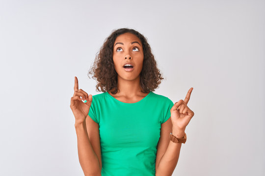 Young brazilian woman wearing green t-shirt standing over isolated white background amazed and surprised looking up and pointing with fingers and raised arms.