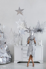 Beautiful and modish young woman with silver bright makeup, in bright silver dress and in a silver mask of deer posing near the alternative silver Christmas tree in the interior
