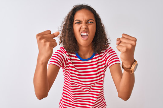 Young brazilian woman wearing red striped t-shirt standing over isolated white background angry and mad raising fists frustrated and furious while shouting with anger. Rage and aggressive concept.