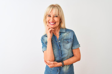 Middle age woman wearing casual denim shirt standing over isolated white background looking confident at the camera smiling with crossed arms and hand raised on chin. Thinking positive.