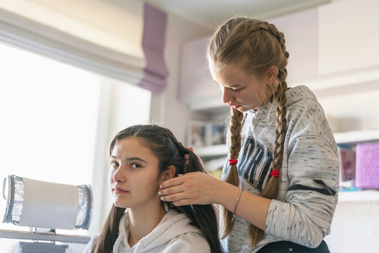 Girl braiding friend's hair in pigtail hairstyle sits on chair in room in front of mirror, window background