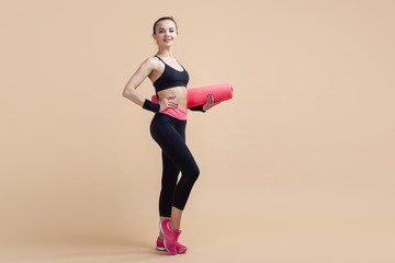 A young girl, a brunette smiling, stands relaxed three-quarters, holding a yoga mat under her arm.