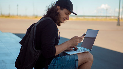 A young brunette man with long hair is sitting on the granite steps in the shade with a laptop and a smartphone.