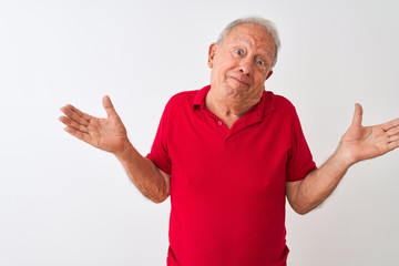 Senior grey-haired man wearing red polo standing over isolated white background clueless and confused expression with arms and hands raised. Doubt concept.