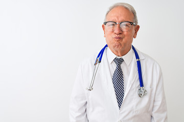 Senior grey-haired doctor man wearing stethoscope standing over isolated white background puffing cheeks with funny face. Mouth inflated with air, crazy expression.