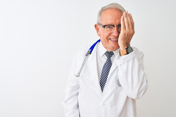 Senior grey-haired doctor man wearing stethoscope standing over isolated white background covering one eye with hand, confident smile on face and surprise emotion.