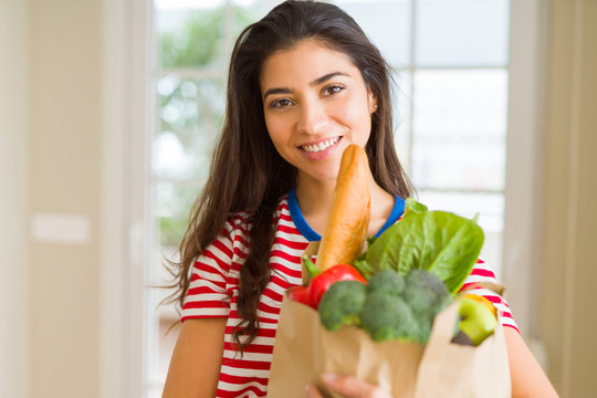Beautiful young woman smiling holding a paper bag full of fresh groceries at home