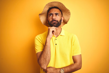 Young indian man on vacation wearing summer hat standing over isolated yellow background with hand on chin thinking about question, pensive expression. Smiling with thoughtful face. Doubt concept.