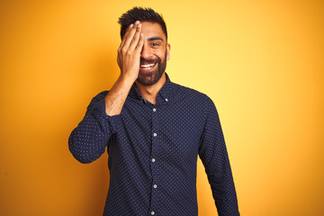 Young handsome indian businessman wearing shirt over isolated yellow background covering one eye with hand, confident smile on face and surprise emotion.