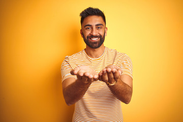 Young indian man wearing t-shirt standing over isolated yellow background Smiling with hands palms...