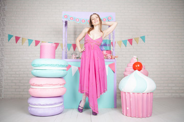 cute happy adult girl posing in fashionable dress among huge fabulous sweets and cakes