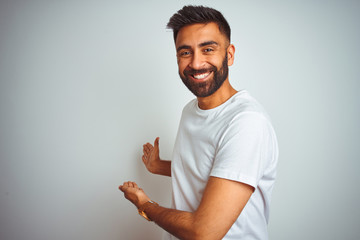 Young indian man wearing t-shirt standing over isolated white background Inviting to enter smiling natural with open hand - obrazy, fototapety, plakaty