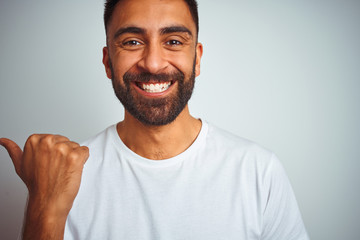 Young indian man wearing t-shirt standing over isolated white background smiling with happy face looking and pointing to the side with thumb up.