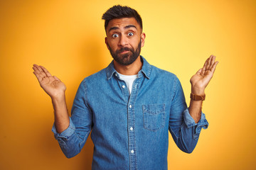 Young indian man wearing denim shirt standing over isolated yellow background clueless and confused...