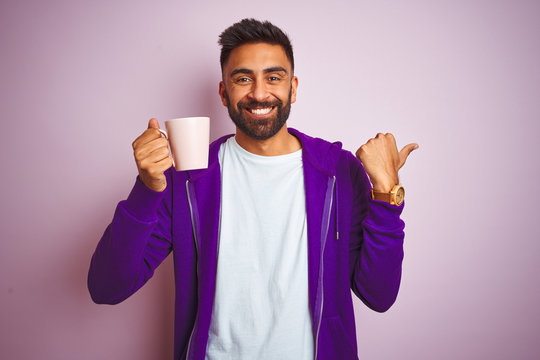 Indian man wearing purple sweatshirt drinking cup of coffee over isolated pink background pointing and showing with thumb up to the side with happy face smiling