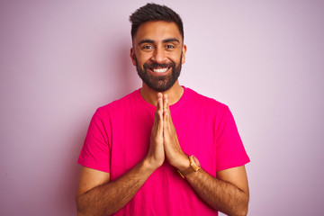 Obraz premium Young indian man wearing t-shirt standing over isolated pink background praying with hands together asking for forgiveness smiling confident.