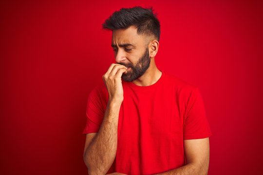 Young handsome indian man wearing t-shirt over isolated red background looking stressed and nervous with hands on mouth biting nails. Anxiety problem.