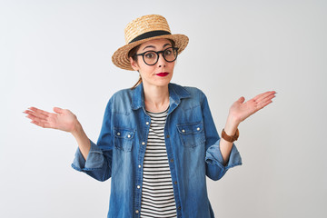 Redhead woman wearing denim shirt glasses and hat over isolated white background clueless and confused expression with arms and hands raised. Doubt concept.