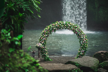 Wedding ceremony on Tibumana waterfall, Bali. Round arch with fresh flowers in jungle. Unusual...