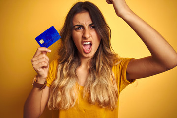 Young beautiful woman holding credit card over yellow isolated background annoyed and frustrated shouting with anger, crazy and yelling with raised hand, anger concept