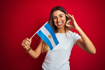 Young beautiful woman holding argentine flag over red isolated background doing ok sign with fingers, excellent symbol