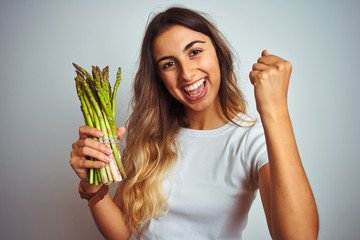 Young beautiful woman eating asparagus over grey isolated background screaming proud and celebrating victory and success very excited, cheering emotion