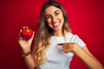 Young beautiful woman holding pepper over red isolated background with surprise face pointing finger to himself