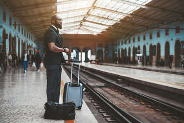 Naklejka na ściany i meble An elegant African businessman is waiting for his train indoors of a railway station depot; a fancy bearded bald Afro senior in eyeglasses with his bags on a railroad platform
