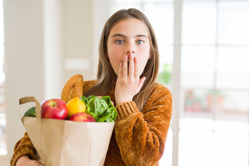 Beautiful young girl holding paper bag of fresh groceries cover mouth with hand shocked with shame for mistake, expression of fear, scared in silence, secret concept