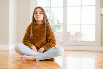 Beautiful young girl kid sitting on the floor at home with serious expression on face. Simple and natural looking at the camera.