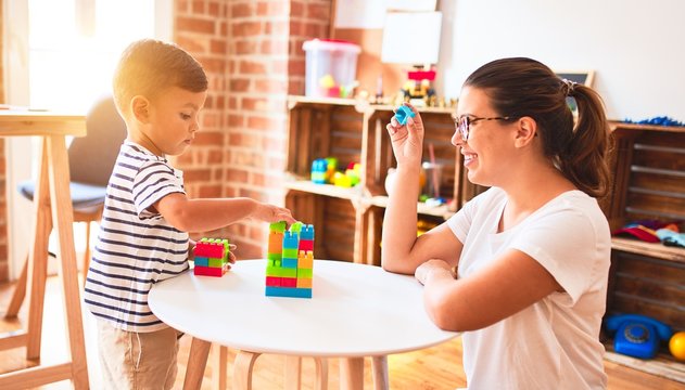 Beautiful teacher and toddler boy playing with construction blocks bulding tower at kindergarten